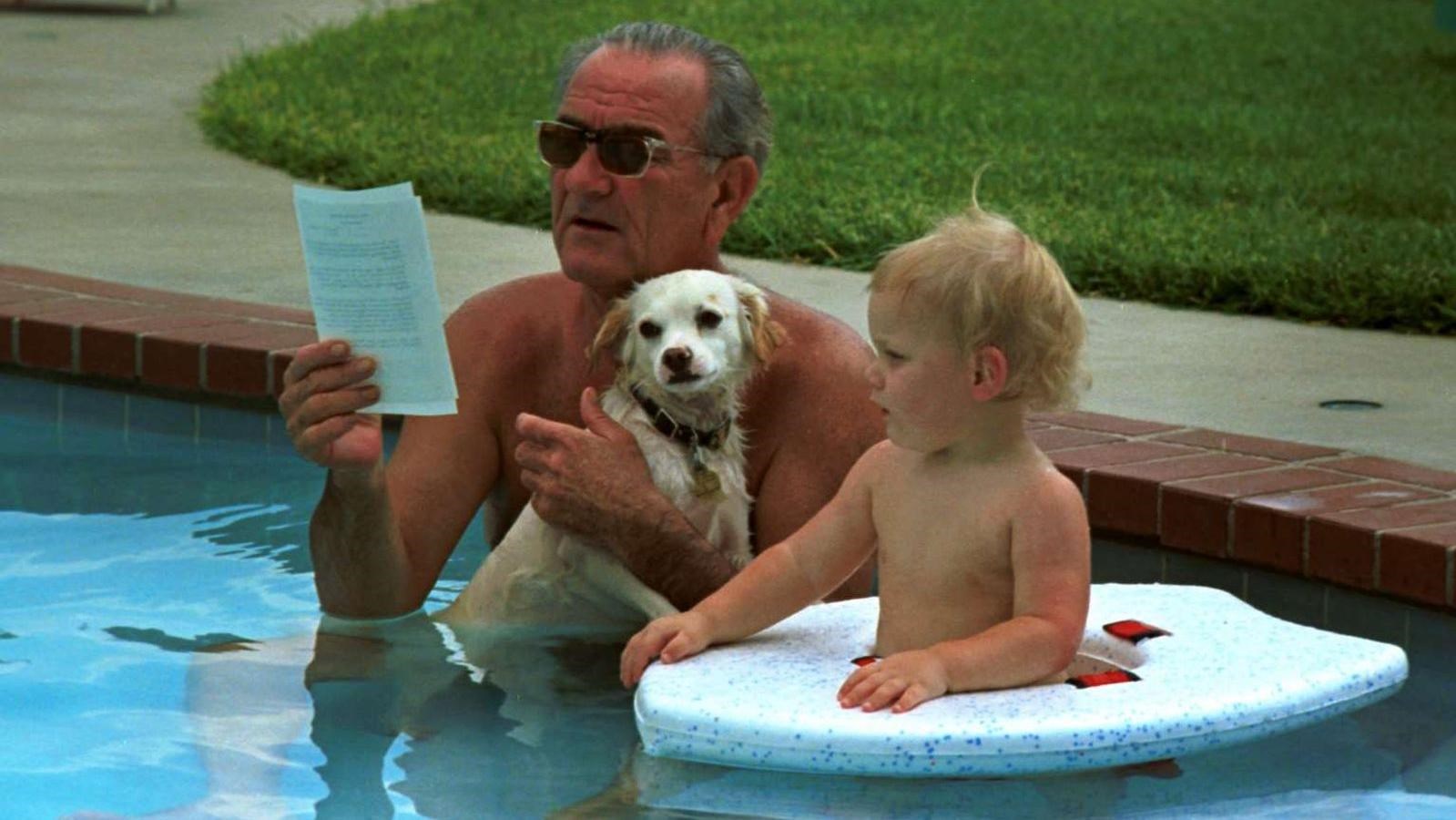 While he reads papers, the president holds a dog in a swimming pool next to his young grandson.
