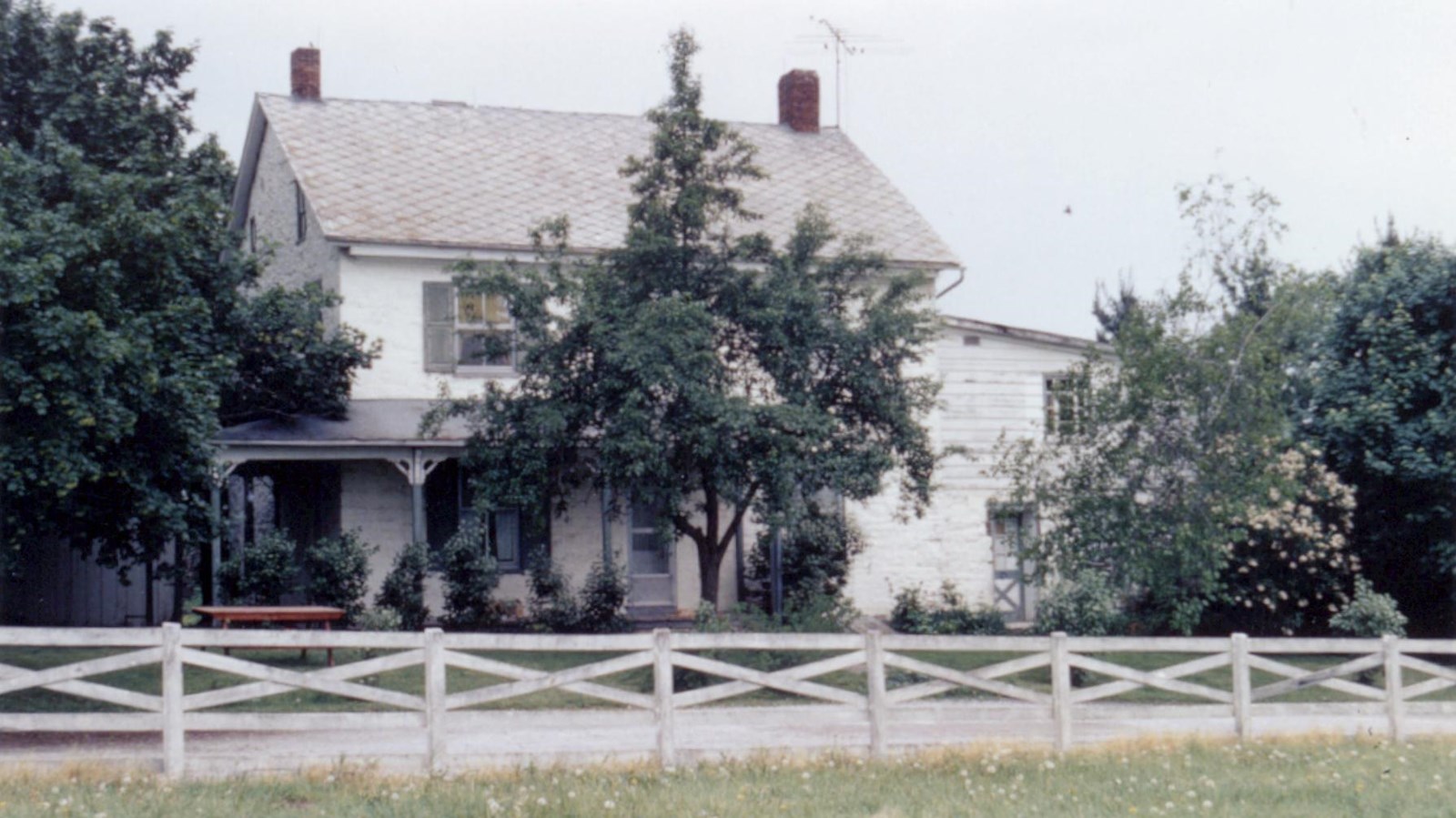 A color image showing a white fence in front of a two-story pale yellow house with multiple trees.