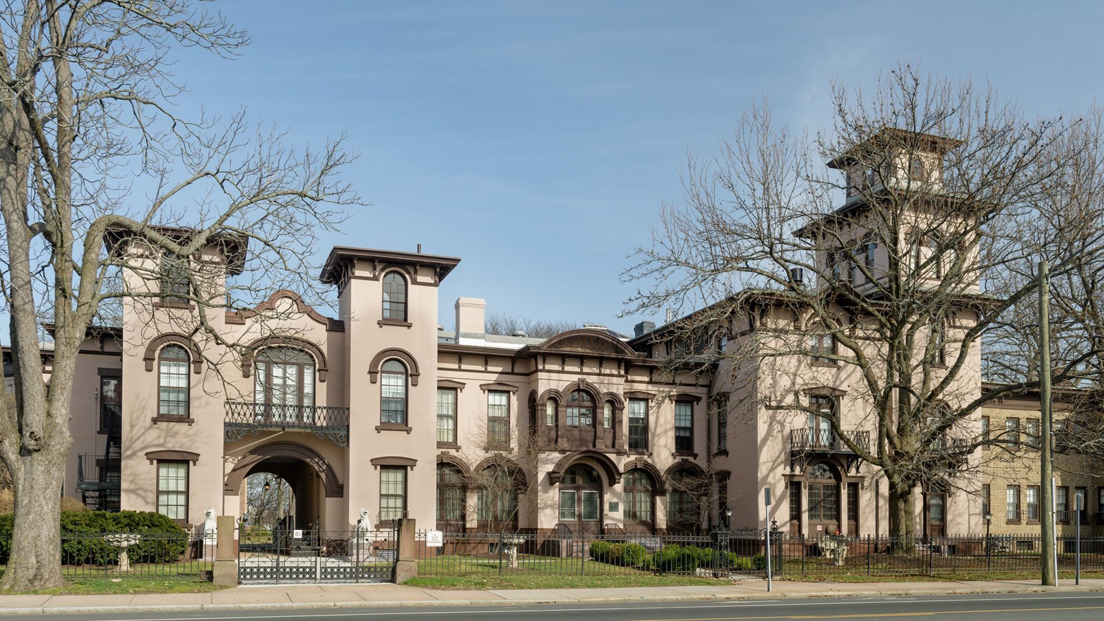 A pink stucco multi-level building, set back from the road with a fence and trees in early spring.