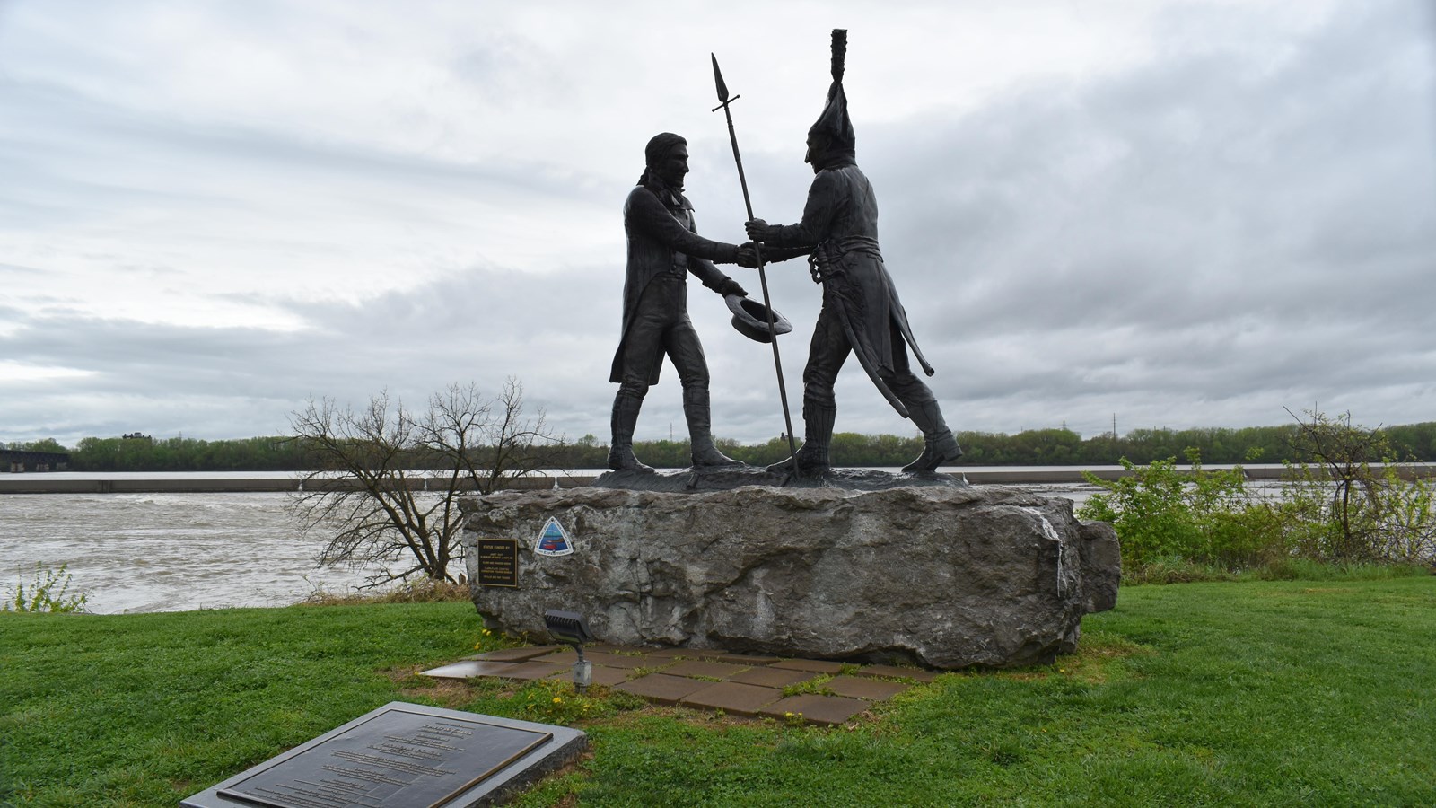 A bronze statue stands atop a large, rough-hewn limestone base beside a wide, fast-moving river unde