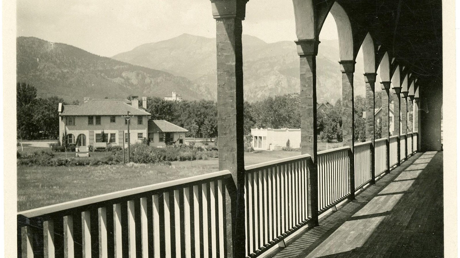Black and white of balcony lined with arches looking out on grassy area, house, and mountains
