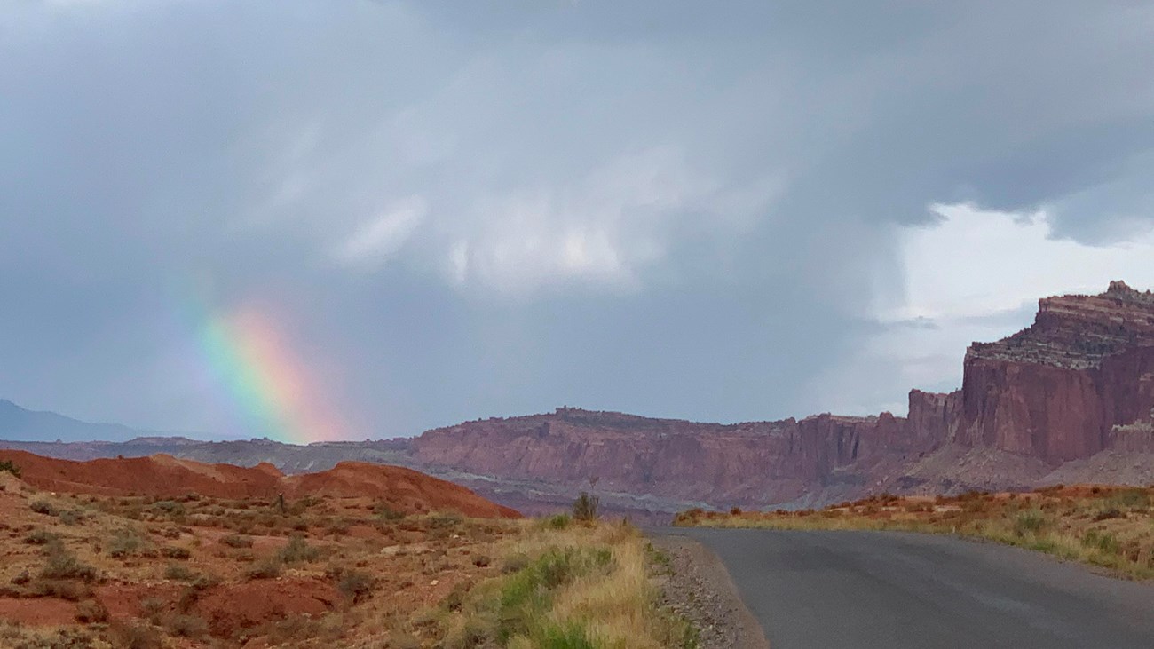 Stormy skies with a small spot of a rainbow, red cliffs, and a blacktop road. 