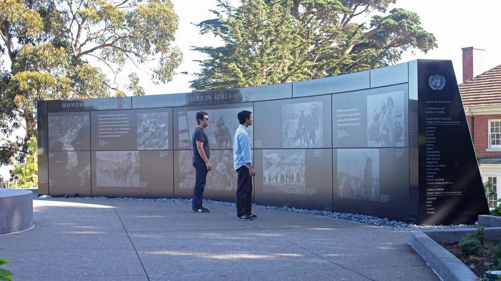 Two men stand in front of a wall commemorating soldiers who served in the Korean War. 