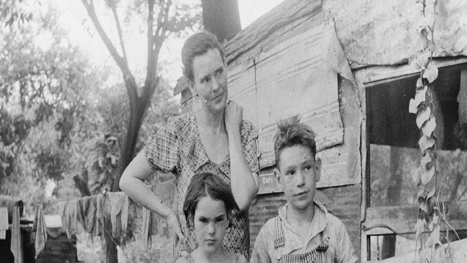 Black and white photo of an unkempt woman and two kids, in the background is a rundown house.n house