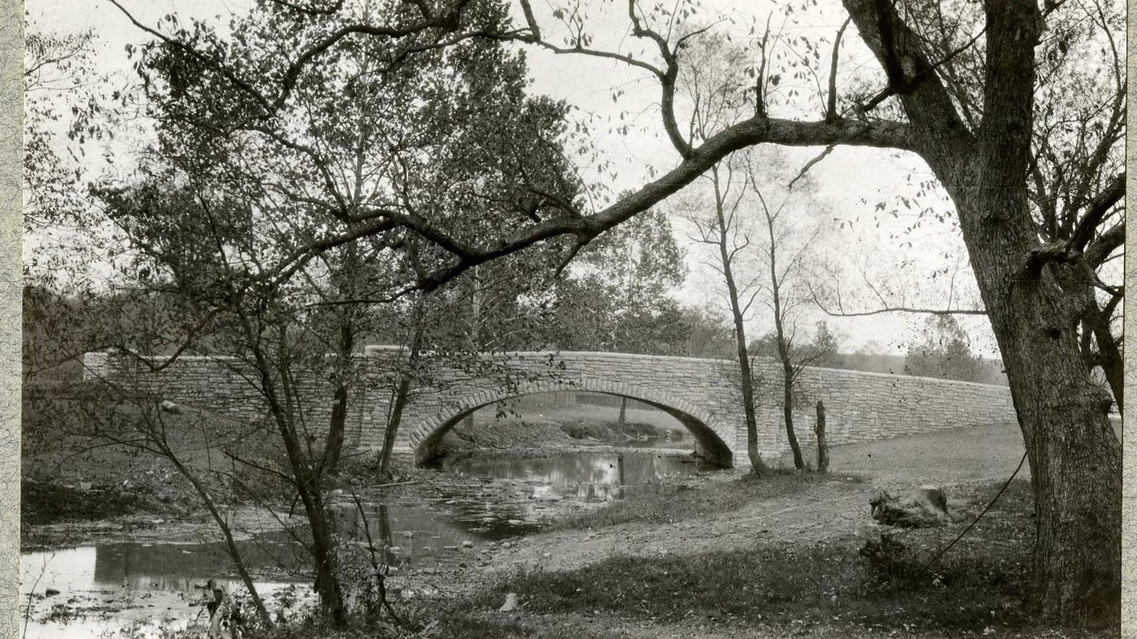 Black and white of stone bridge over water with grass and trees on both sides 
