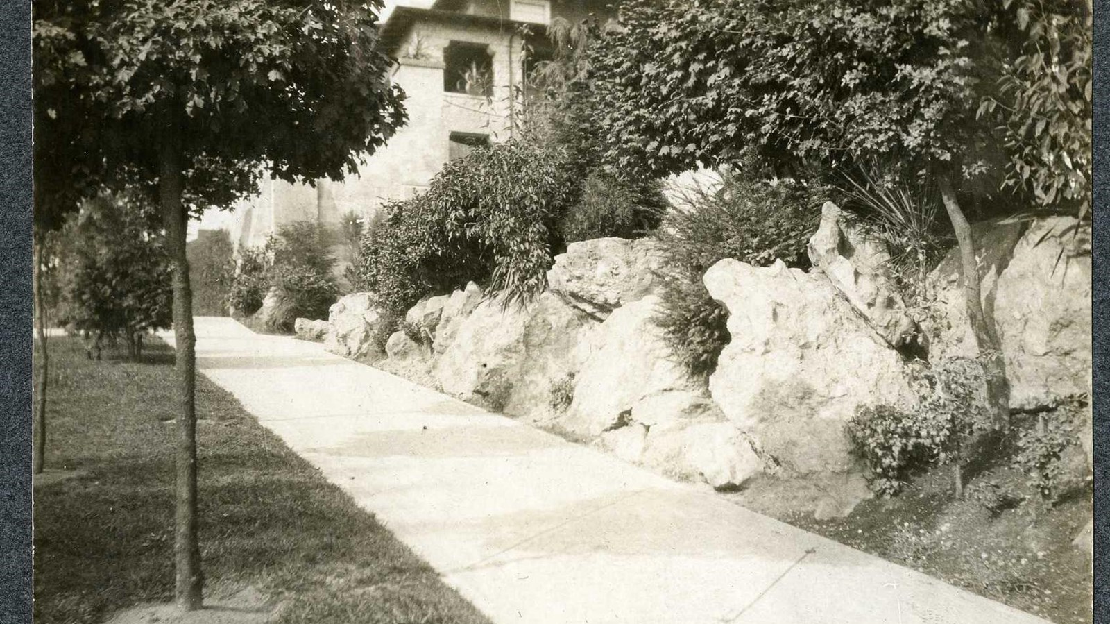 Black and white of sidewalk with grass on one side, rocks on the other and building in distance