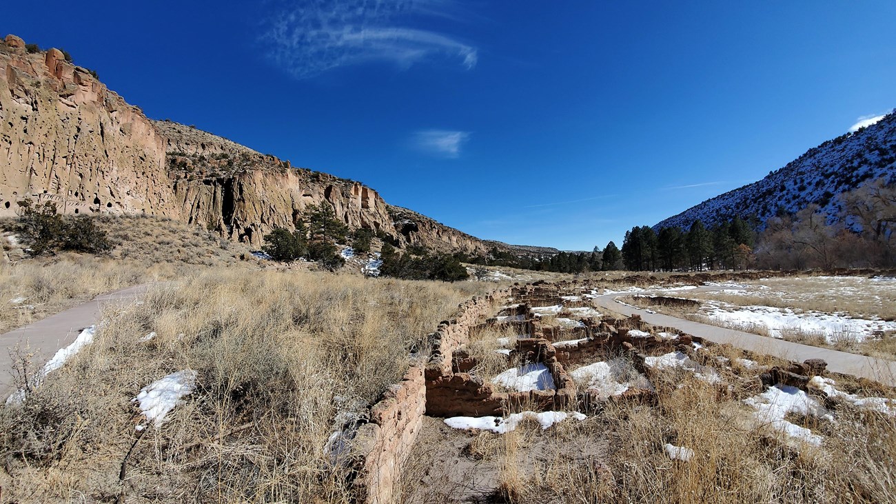 remnants of a stone village in the bottom of a canyon with steep rocky tan cliffs and blue sky