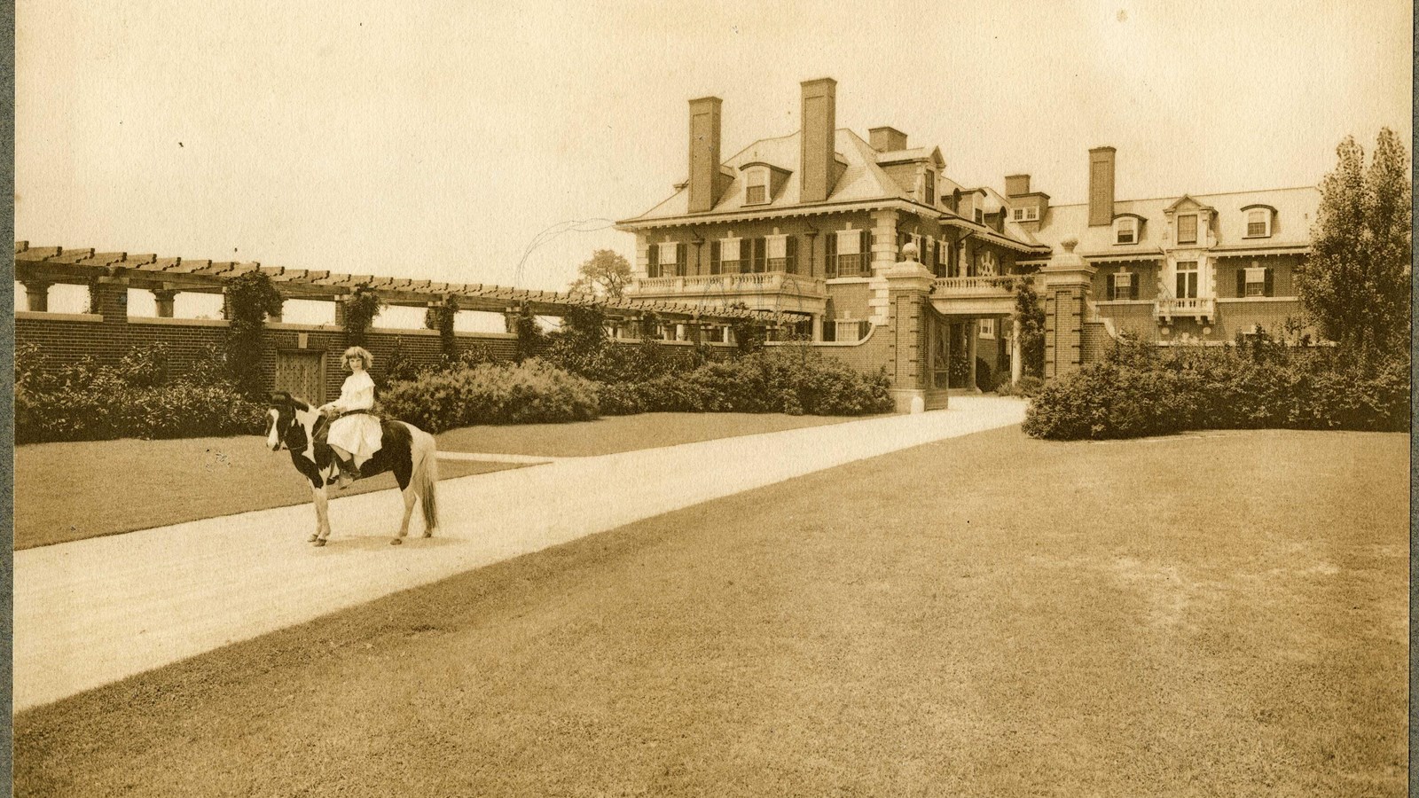 Black and white of flat path with girl riding horse on it, grass on both sides and large home behind