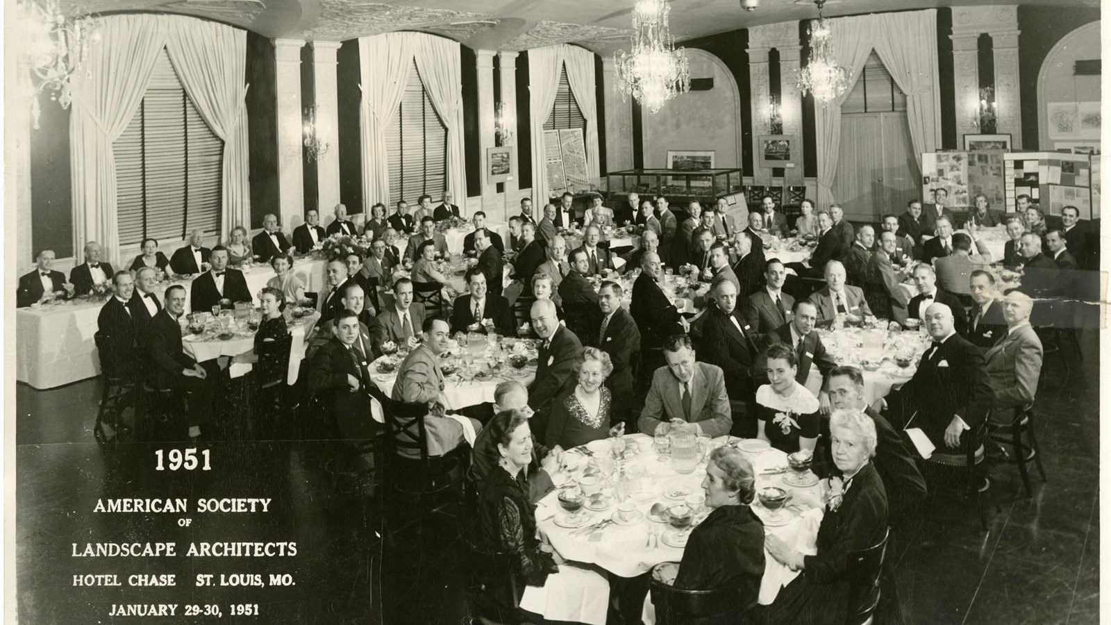 Black and white of hotel room with many tables with people in suits and dresses sitting at the table