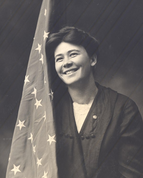 A woman stands smiling facing the viewer holding a large American flag, black and white photo.