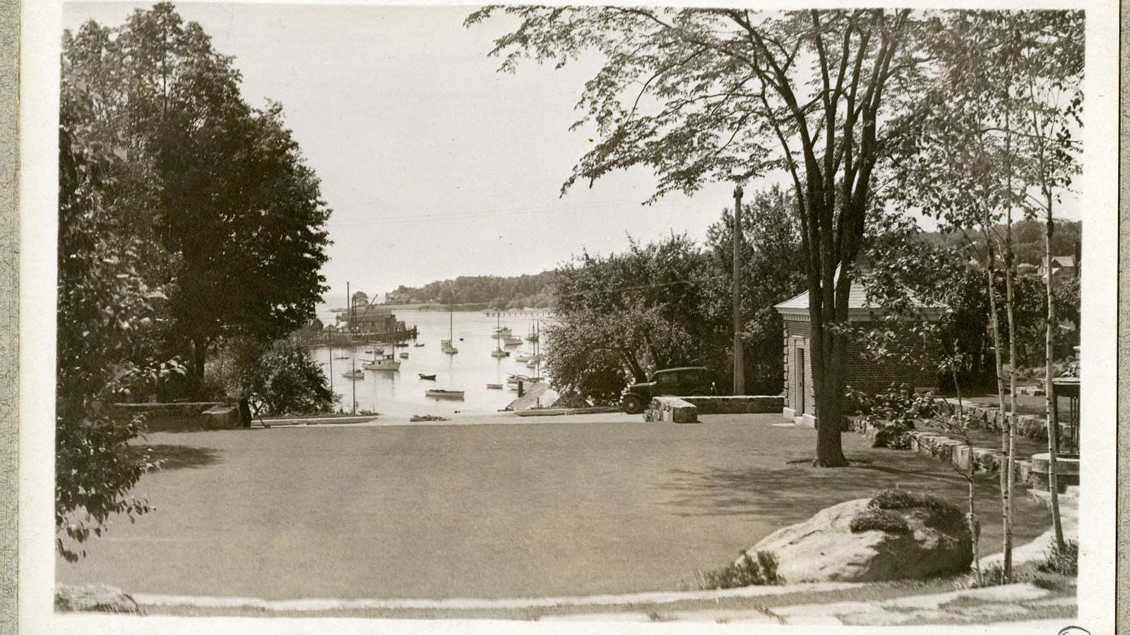 Black and white of flat grassy area with trees on edges overlooking harbor with boats on it