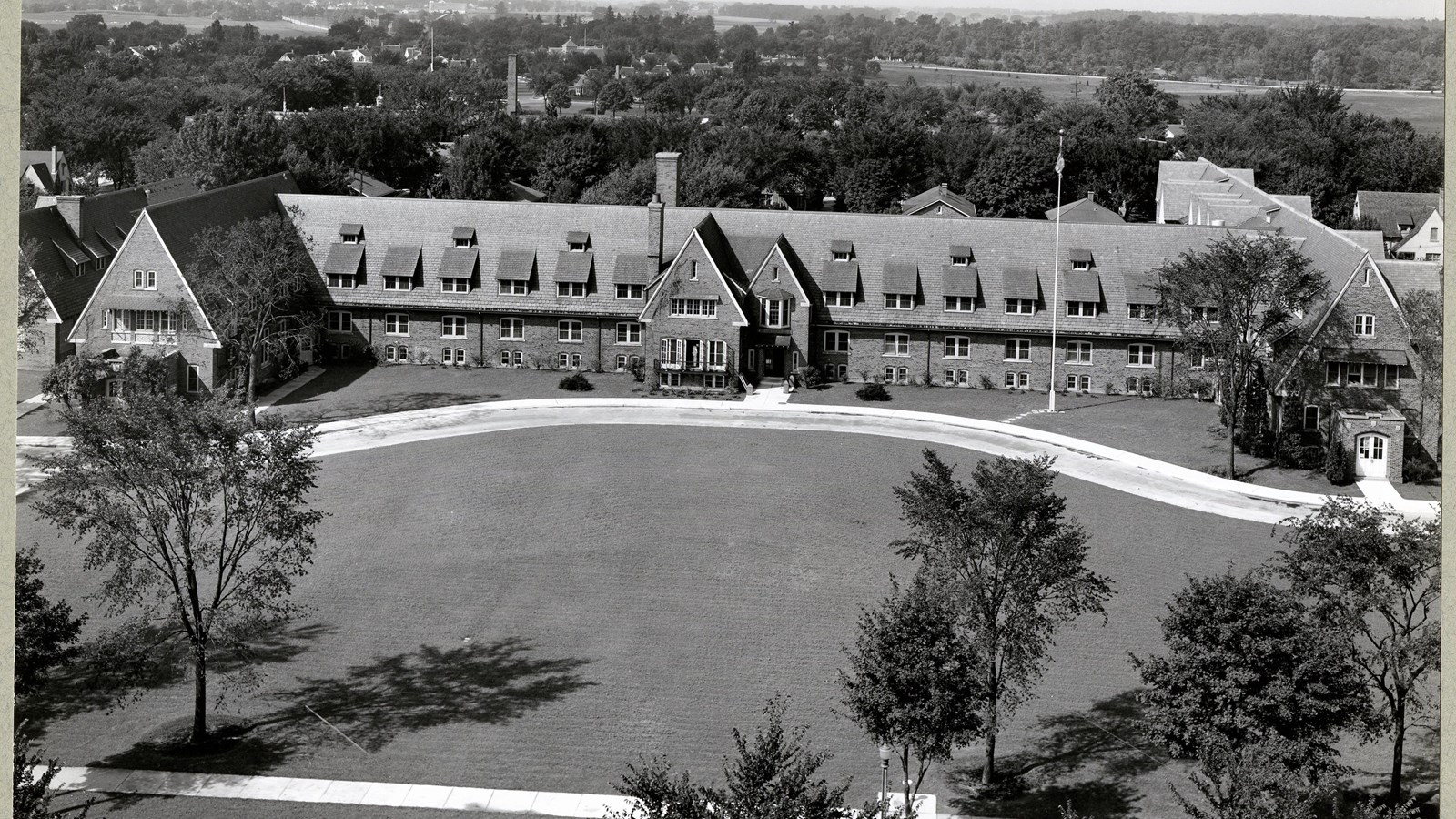 Black and white aerial of large building with grassy area in front, trees around