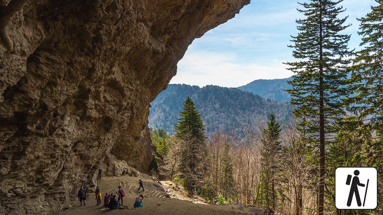 People underneath a rocky overhang with trees and mountains in the background. Hiker icon in corner.