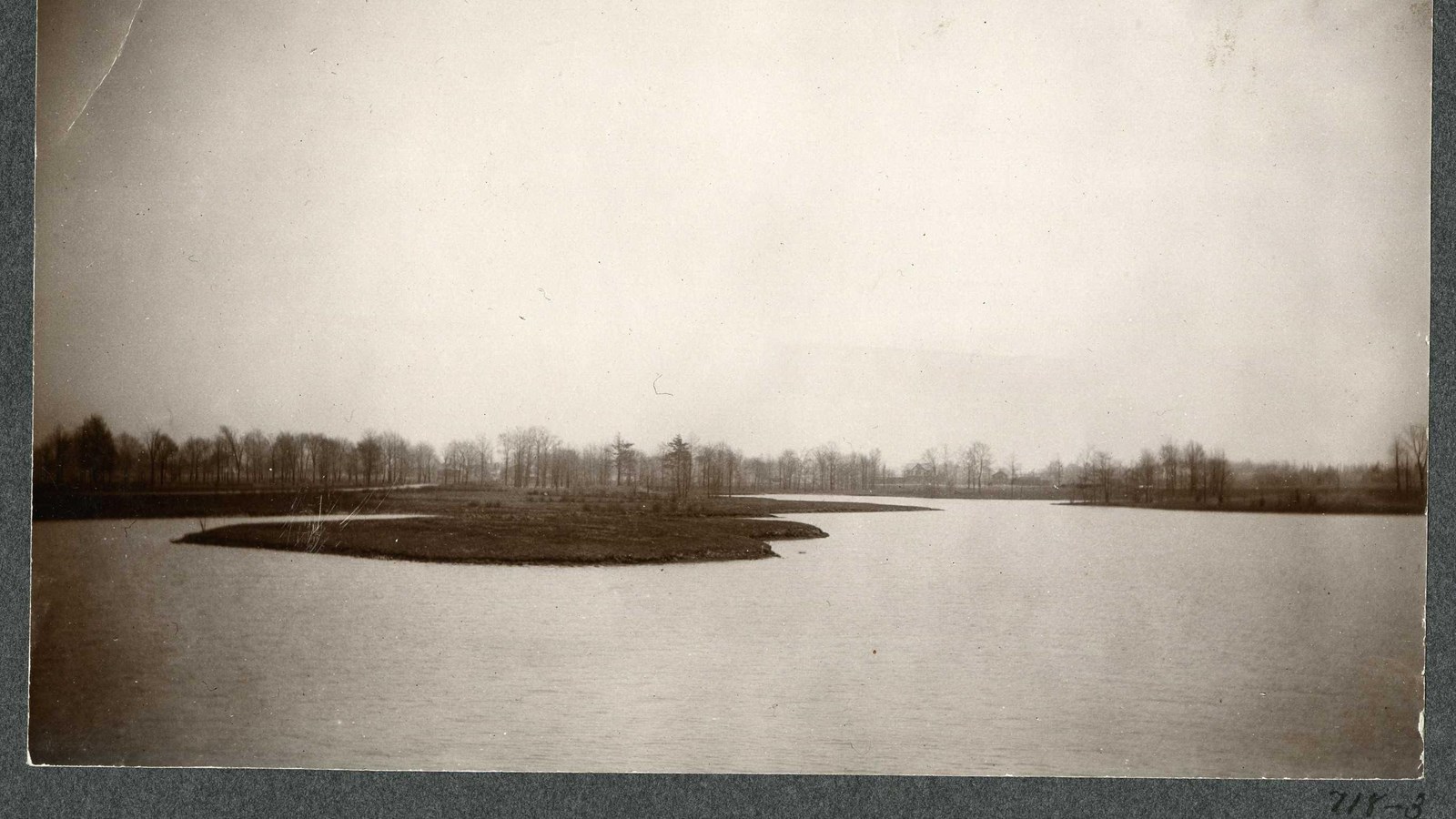 Black and white of flat body of water with flat grassy area jutting into water, trees in distance 