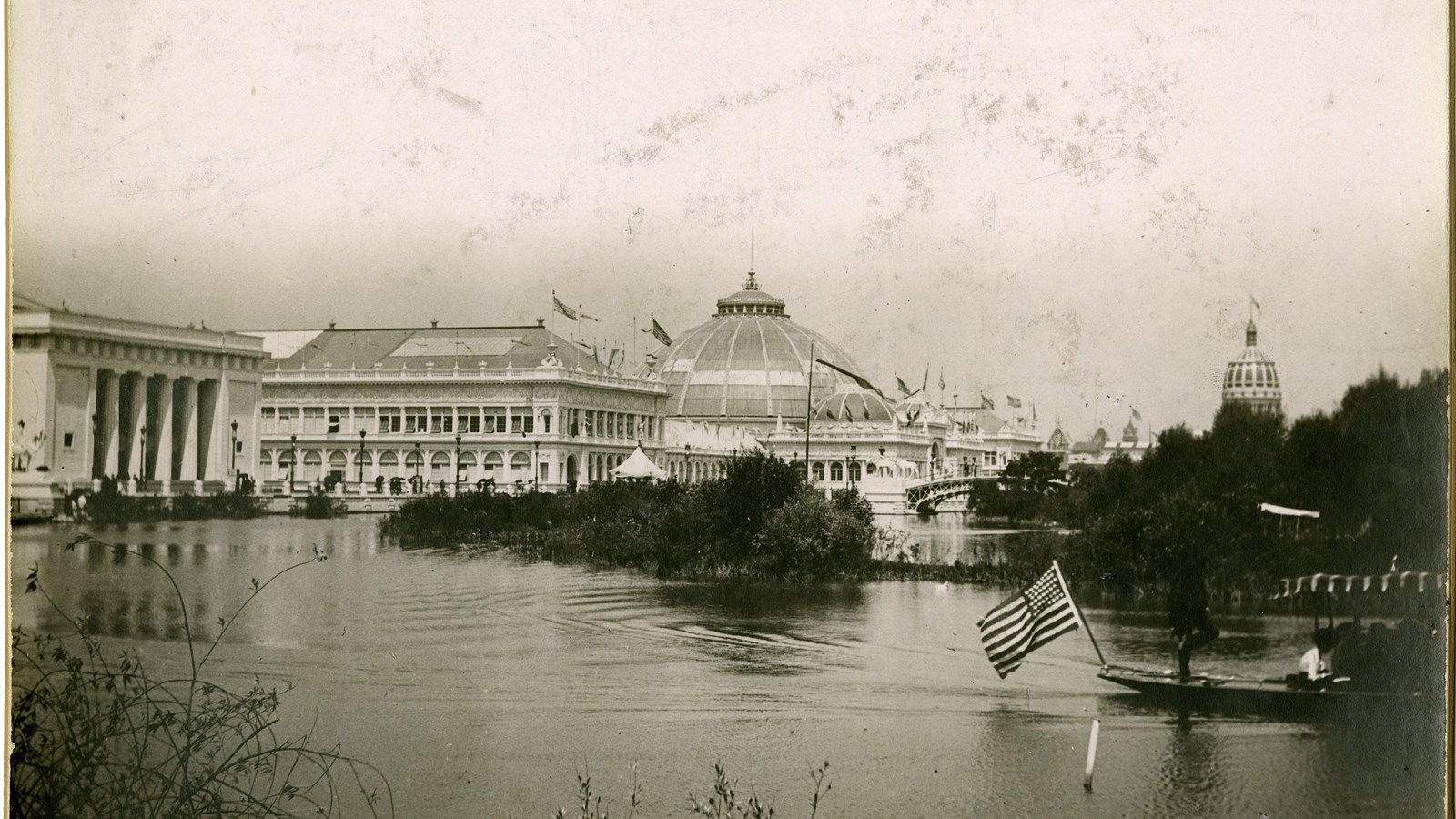 Black and white of body of water with canoe on it with U.S. flag behind, white buildings behind 