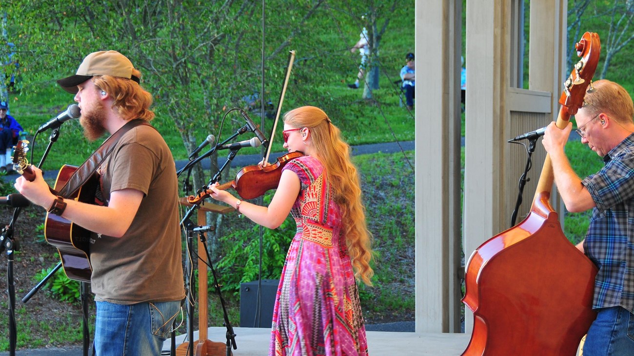 Three musicians with fiddle, bass, and guitar, play on an outdoor stage