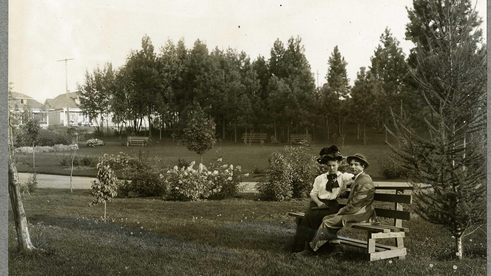 Black and white of bench with two people on flat grassy area with path lined with shrubs