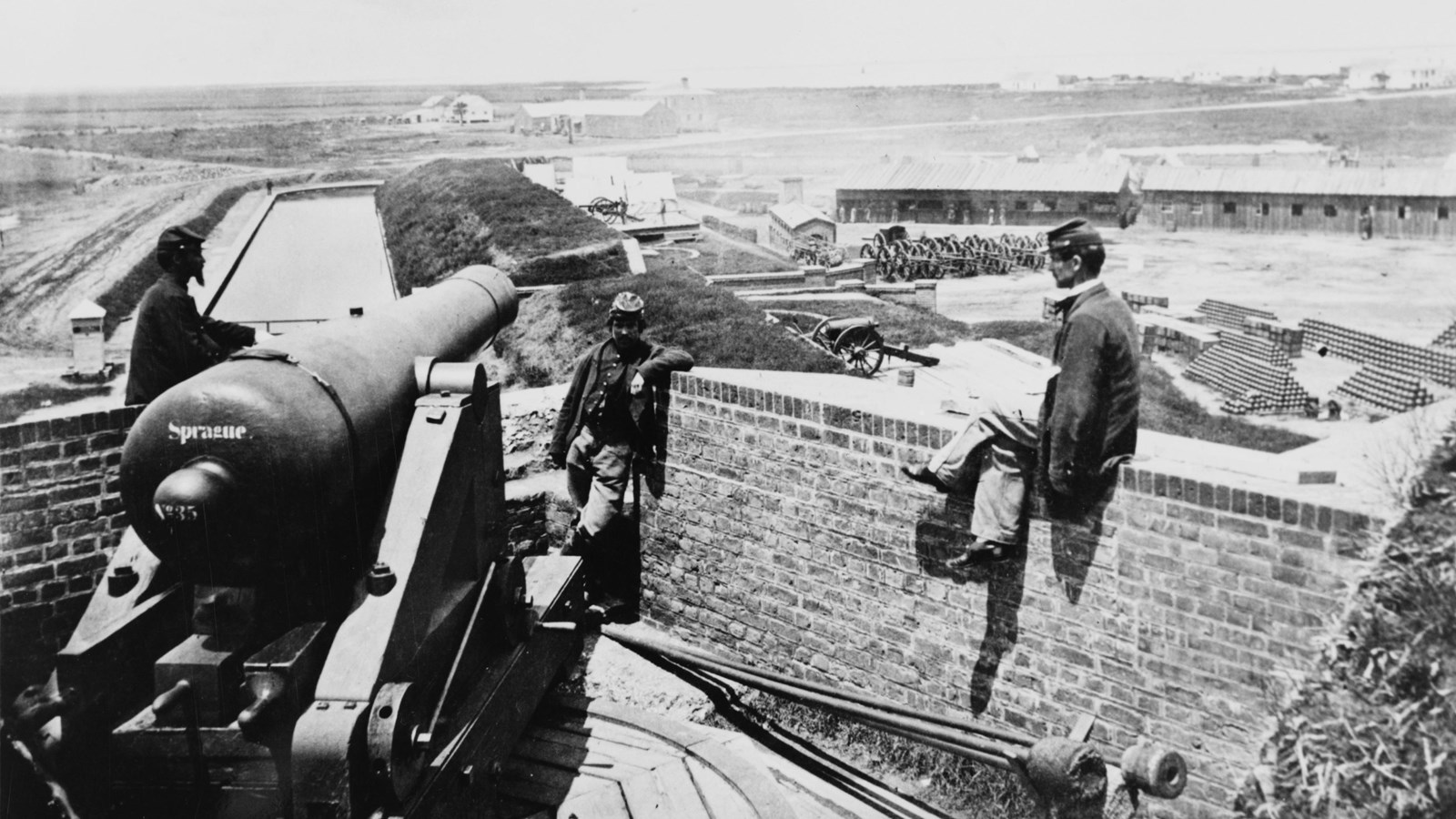 A man sits on a brick wall next to a cannon, several buildings are in the background.
