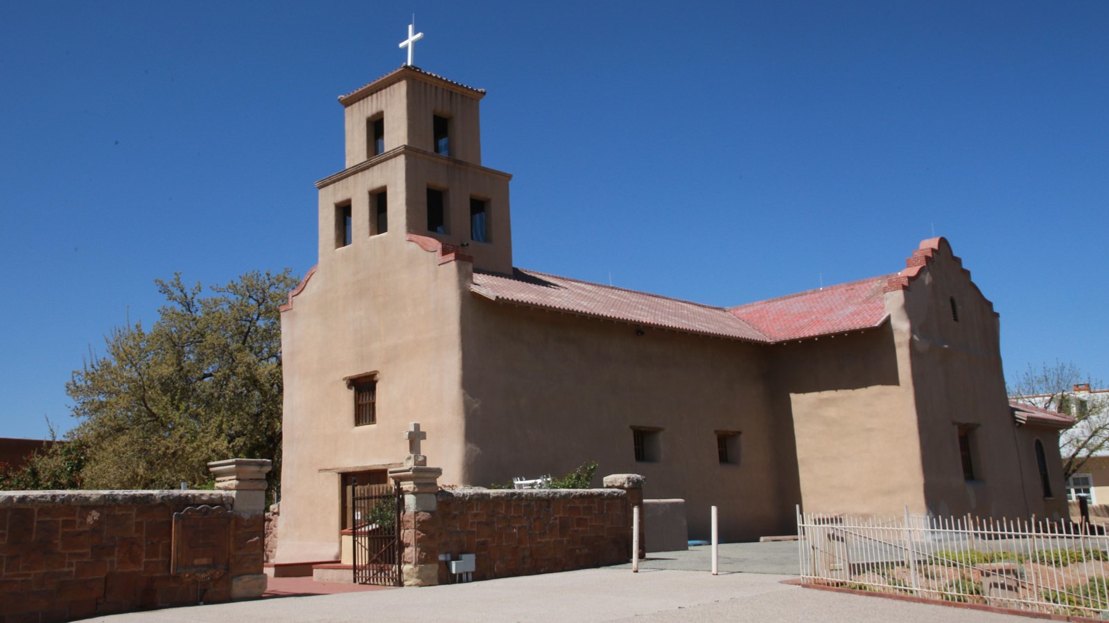 A large, single bell-towered, adobe chapel with a large stone wall surrounding it.