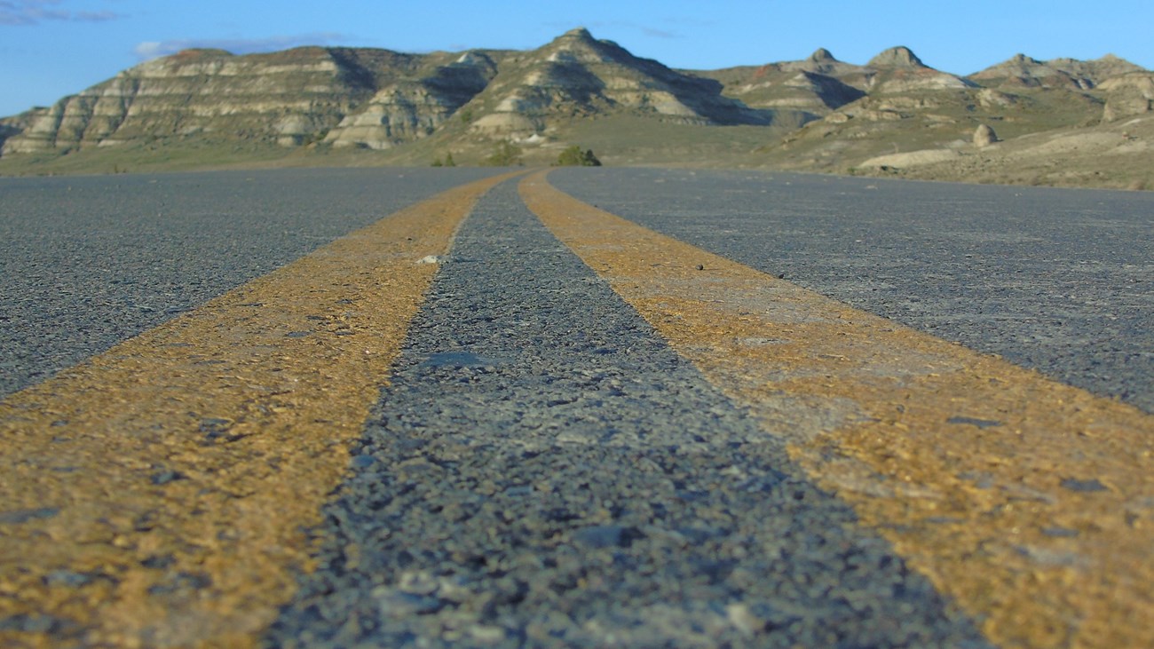 A photo taken from road level. Two yellow lines travel out of sight, with buttes in the distance.