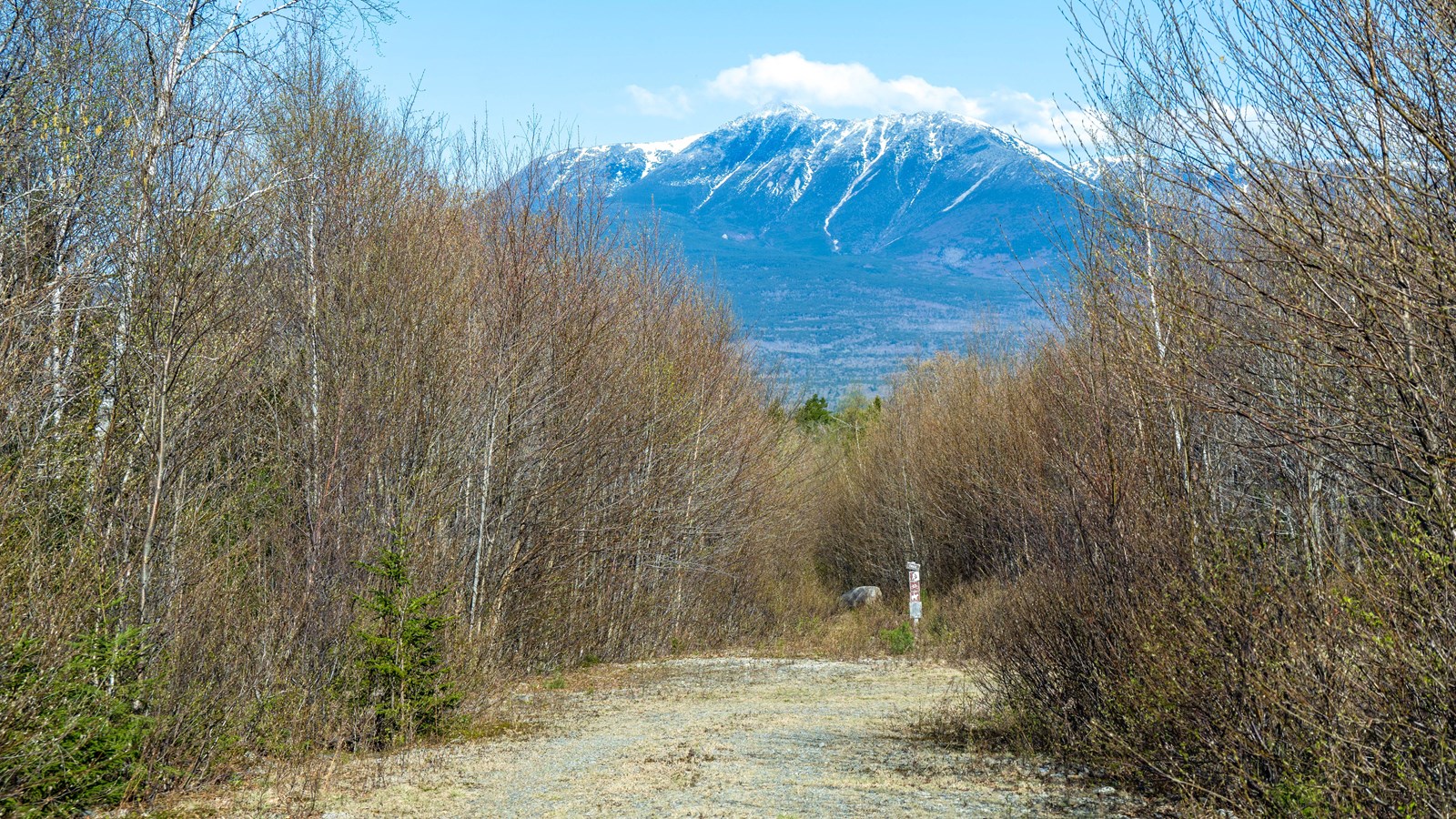 A gravel road leads to a trailhead. A clear view of Katahdin is in the background on a sunny day.
