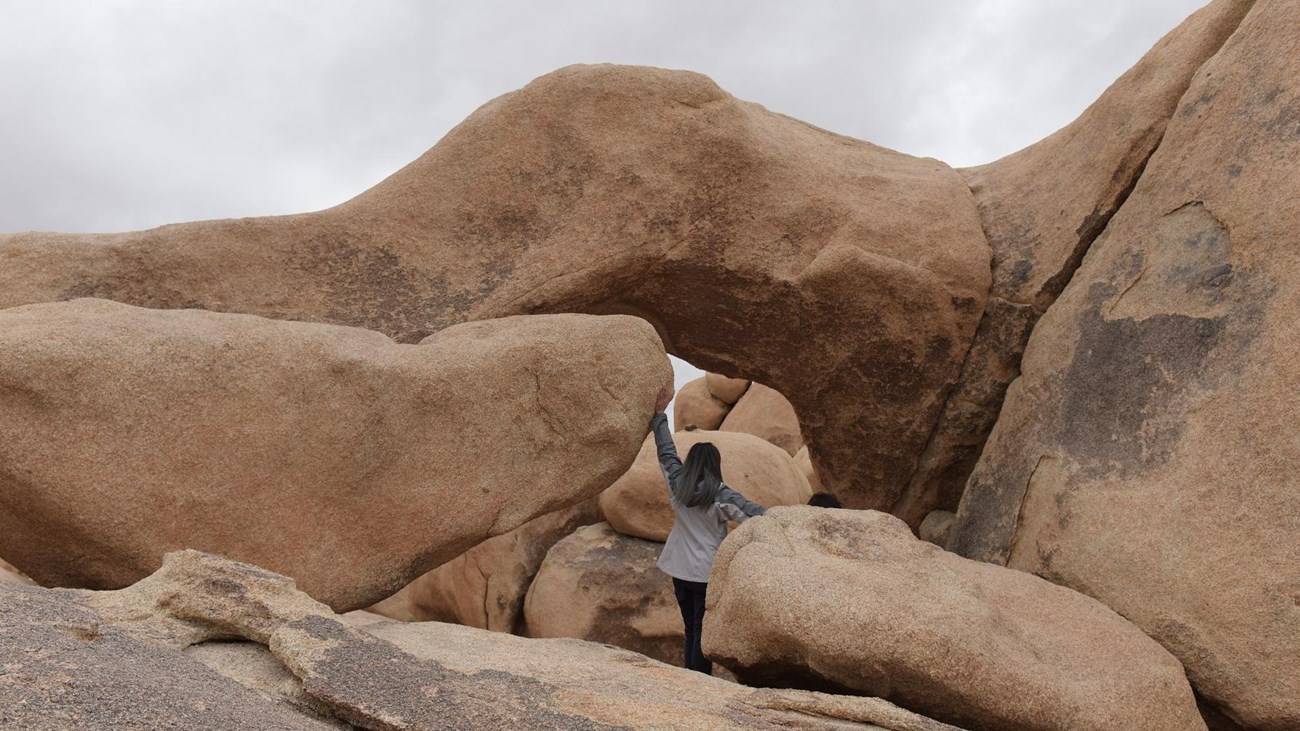 A hiker walking along a barren trail with desert vegetation and small boulders. 