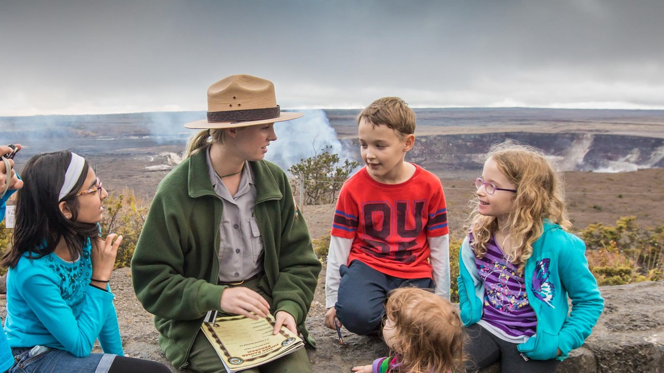 A park ranger sits on a stone wall on the edge of a volcanic caldera surrounded by children