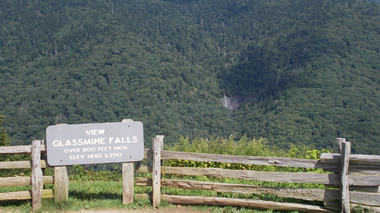 View of a mountain and a tall waterfall with overlook sign and wood fence in the foreground