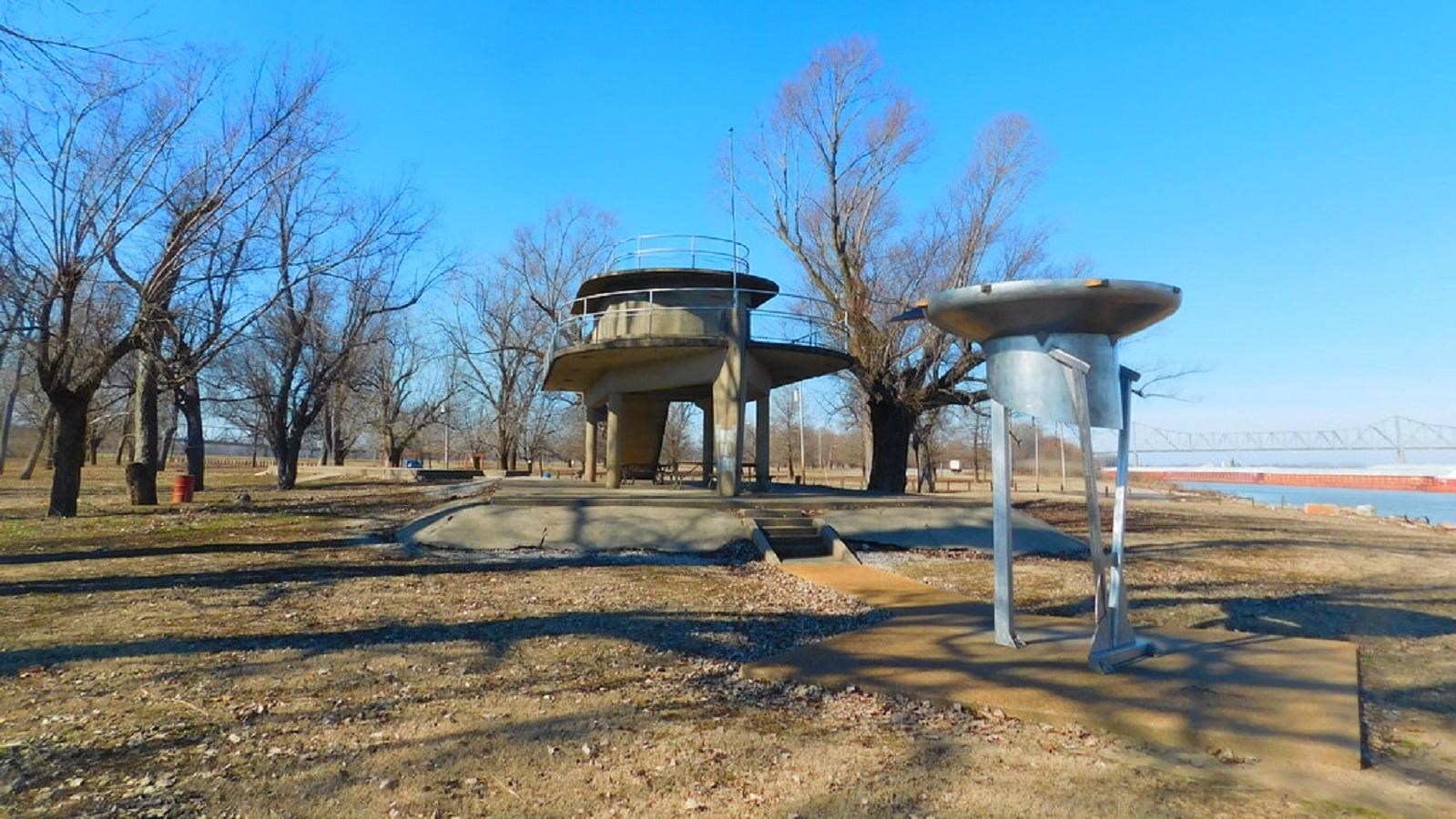 A tall stone observation station and metal sculpture stand along a lake shore