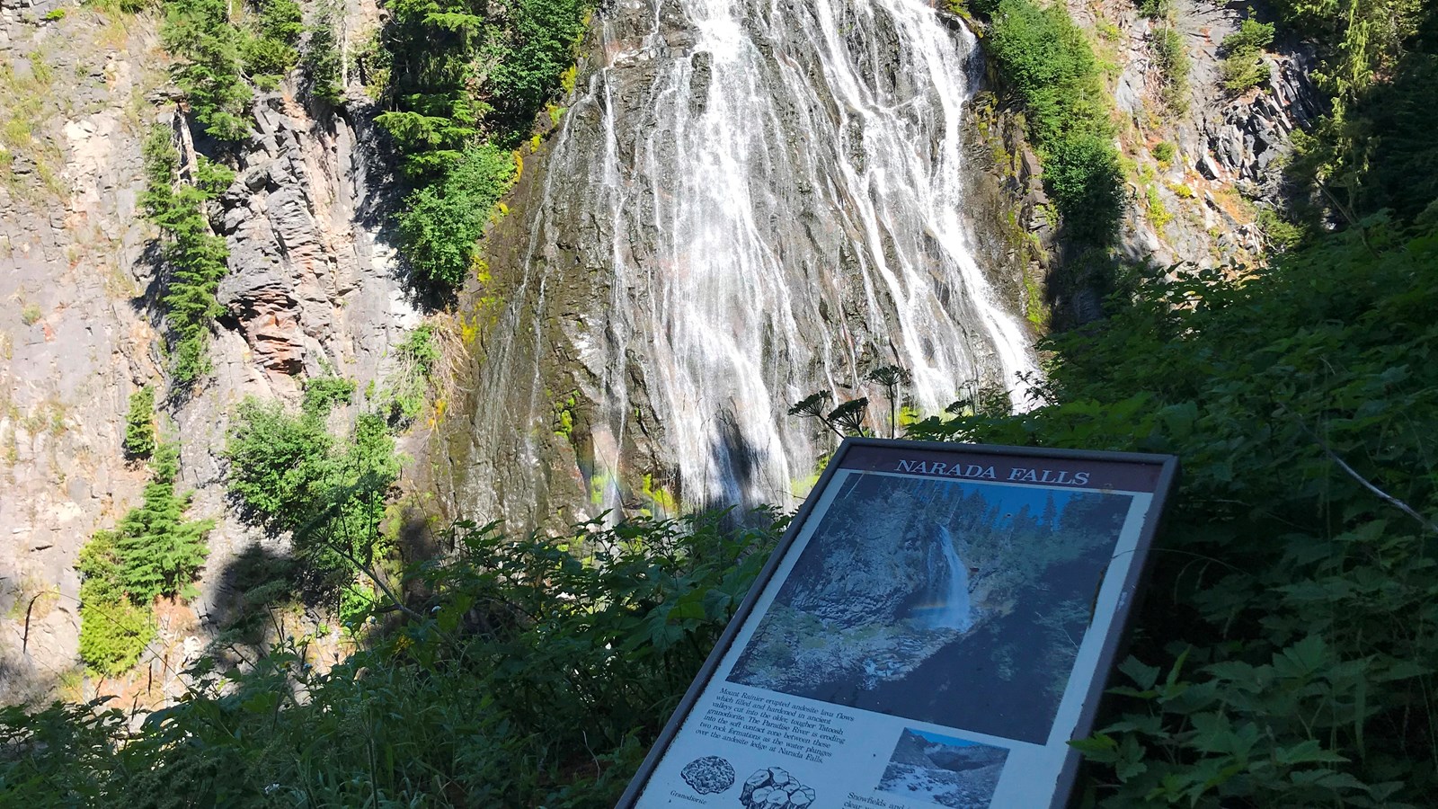 A small panel next to a fence across a narrow gorge from a wide fan-shaped waterfall.