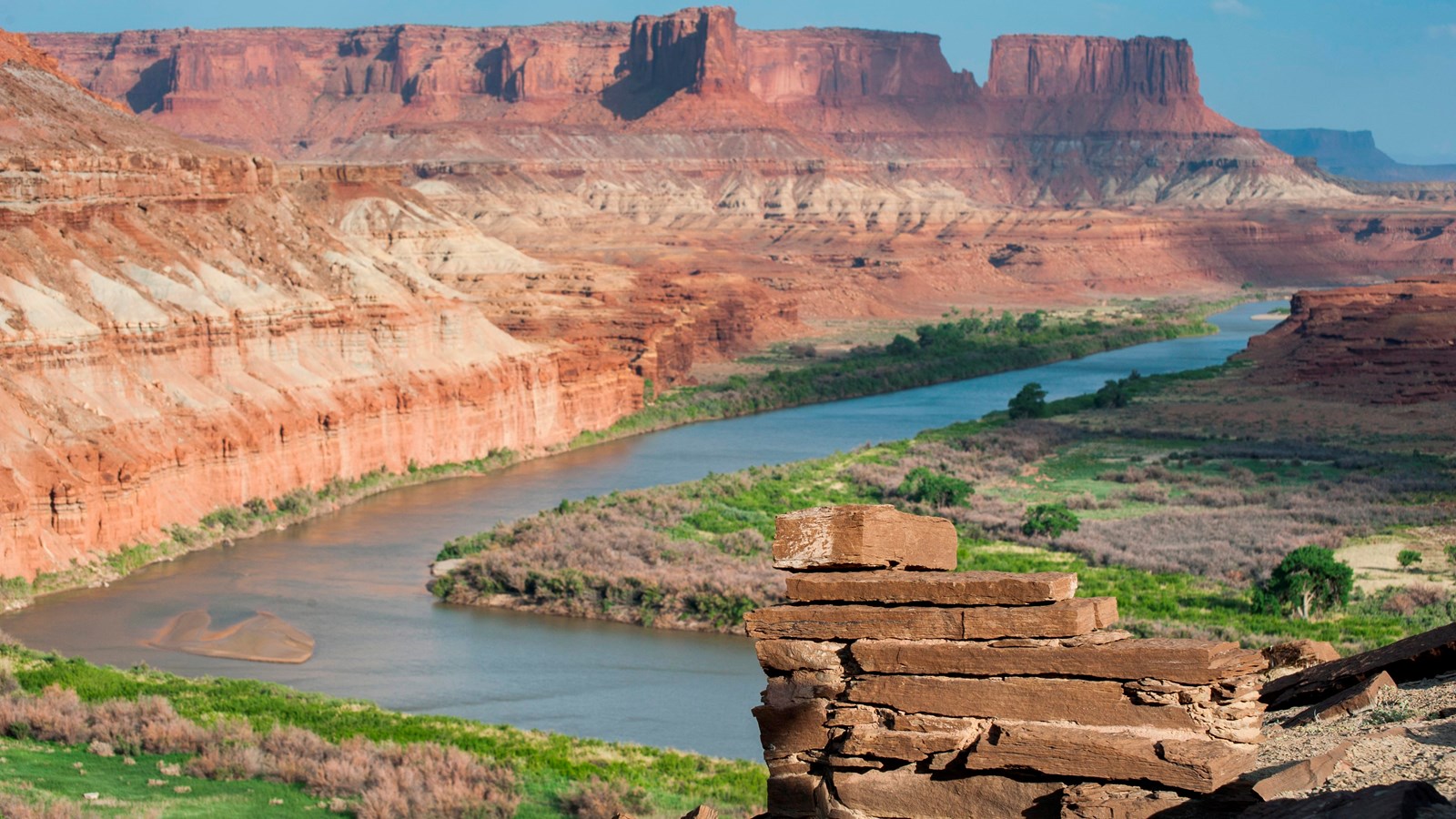 Stacked sandstone slabs sit atop a cliff overlooking the green river.