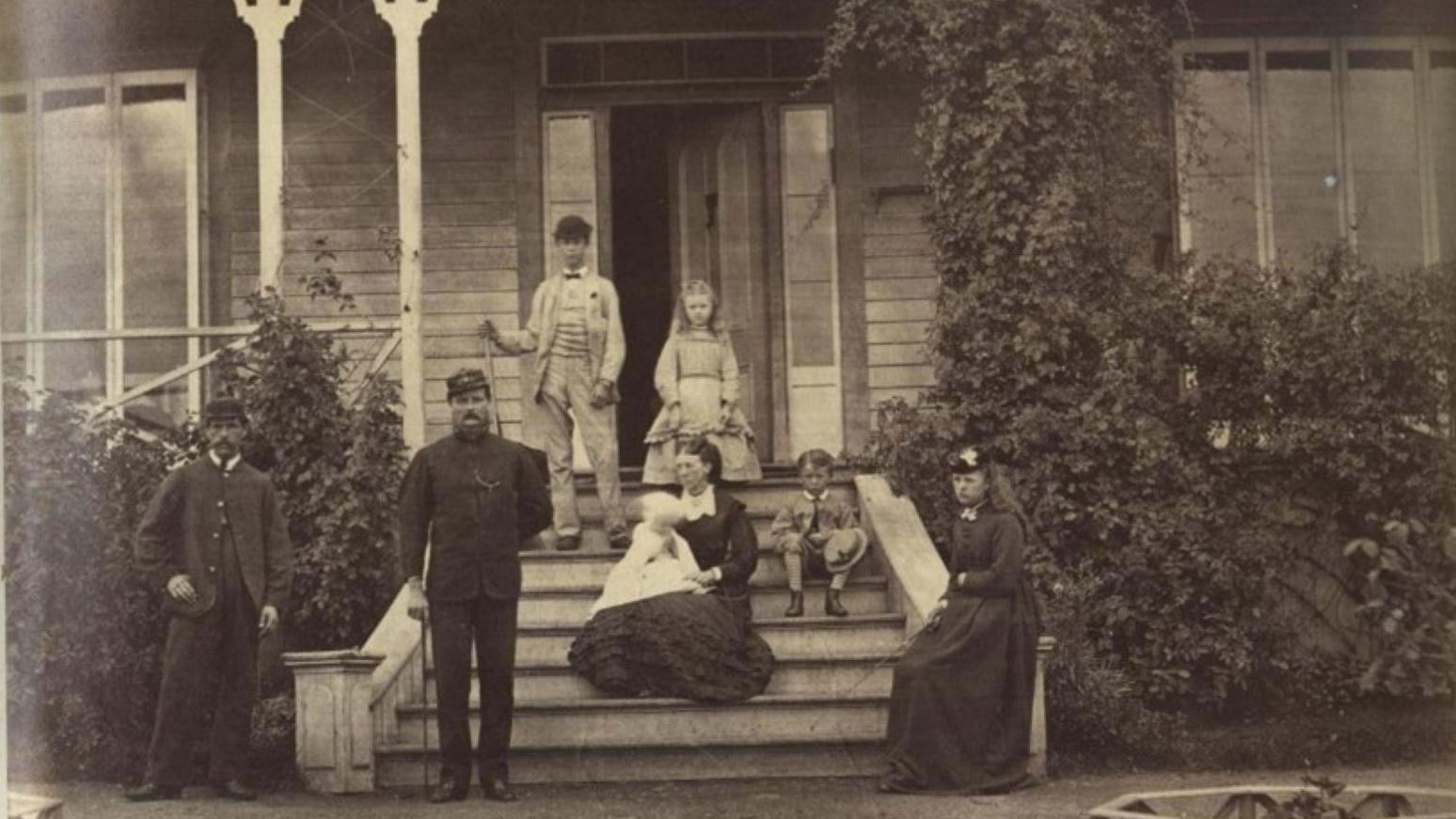 Black and white photograph of a Victorian family sitting on the steps of an ivy covered cottage.