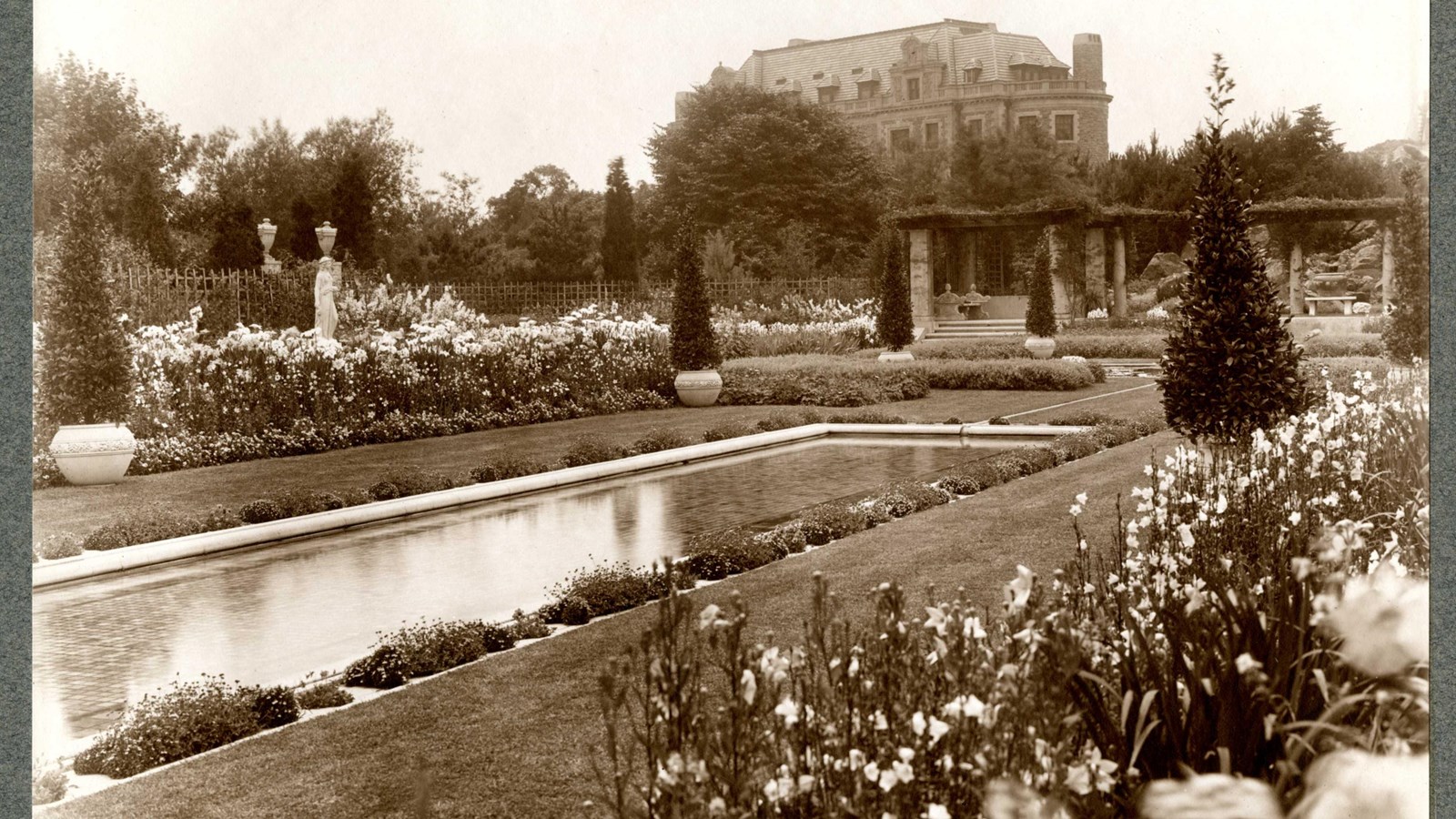 Black and white of rectangular pool on flat grassy area with flowers on edge, large house on hill