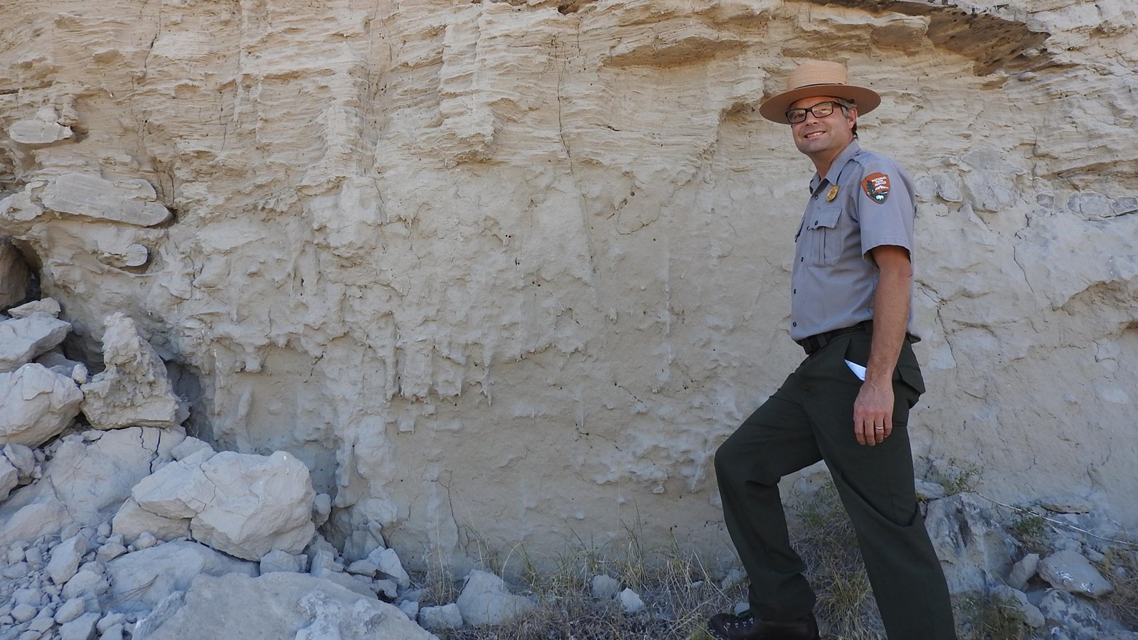 A park ranger stands near an area where stratified sandstone is seen above massive sandstone.