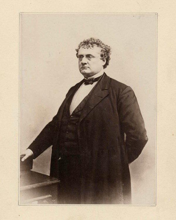 White man with short curly hair & glasses, wearing a dark three-piece suit standing next to a desk. 