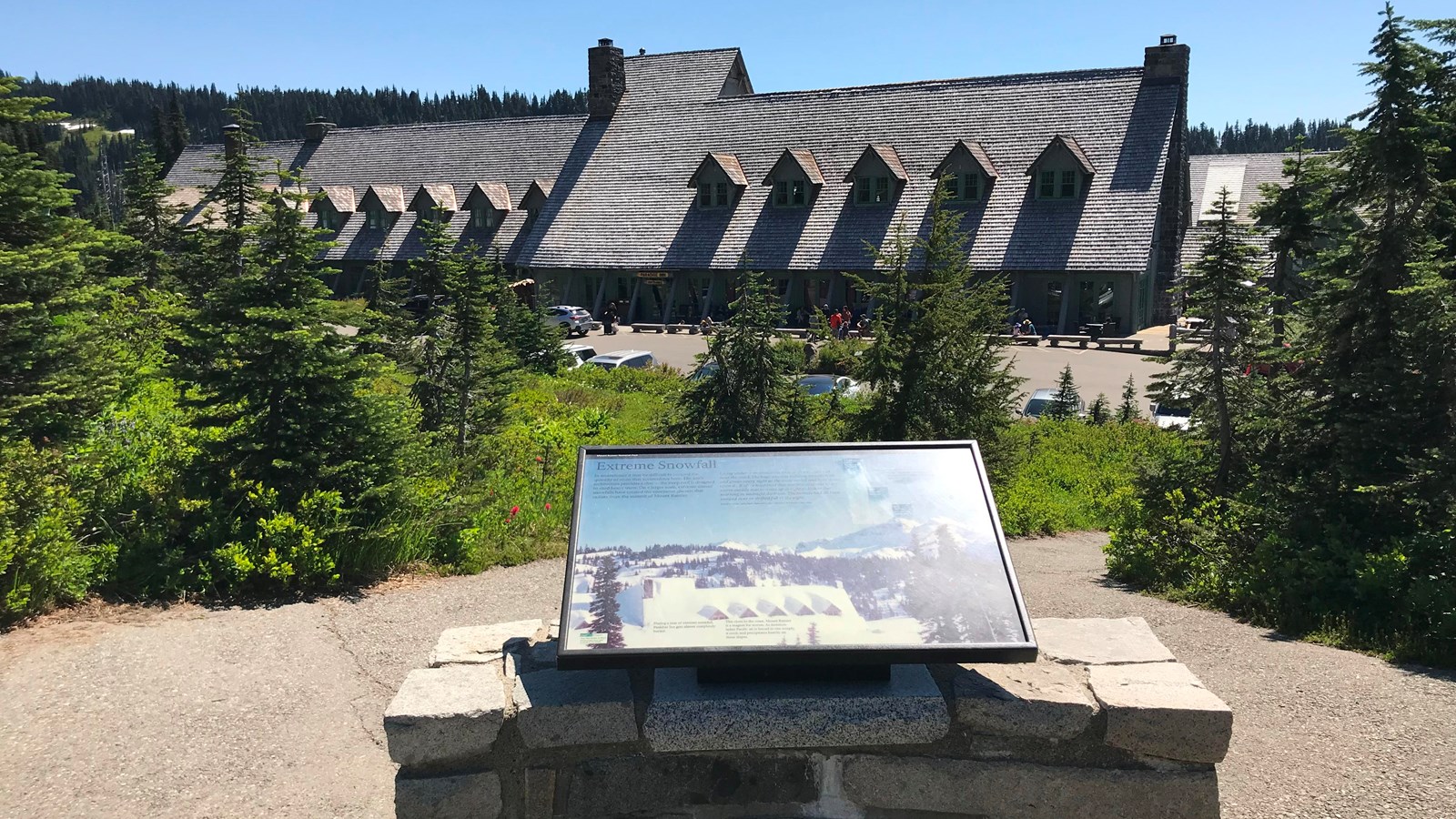 A wayside panel attached to rock wall overlooking a large wood building with a steep roof. 