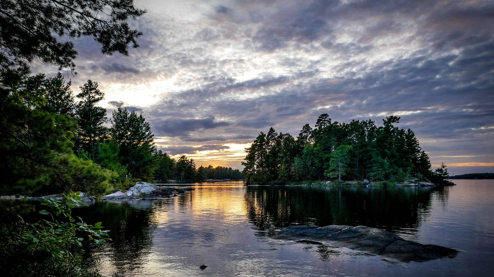 A sunset view on a lake with reflections in the water. An island is in view that has a dense forest.