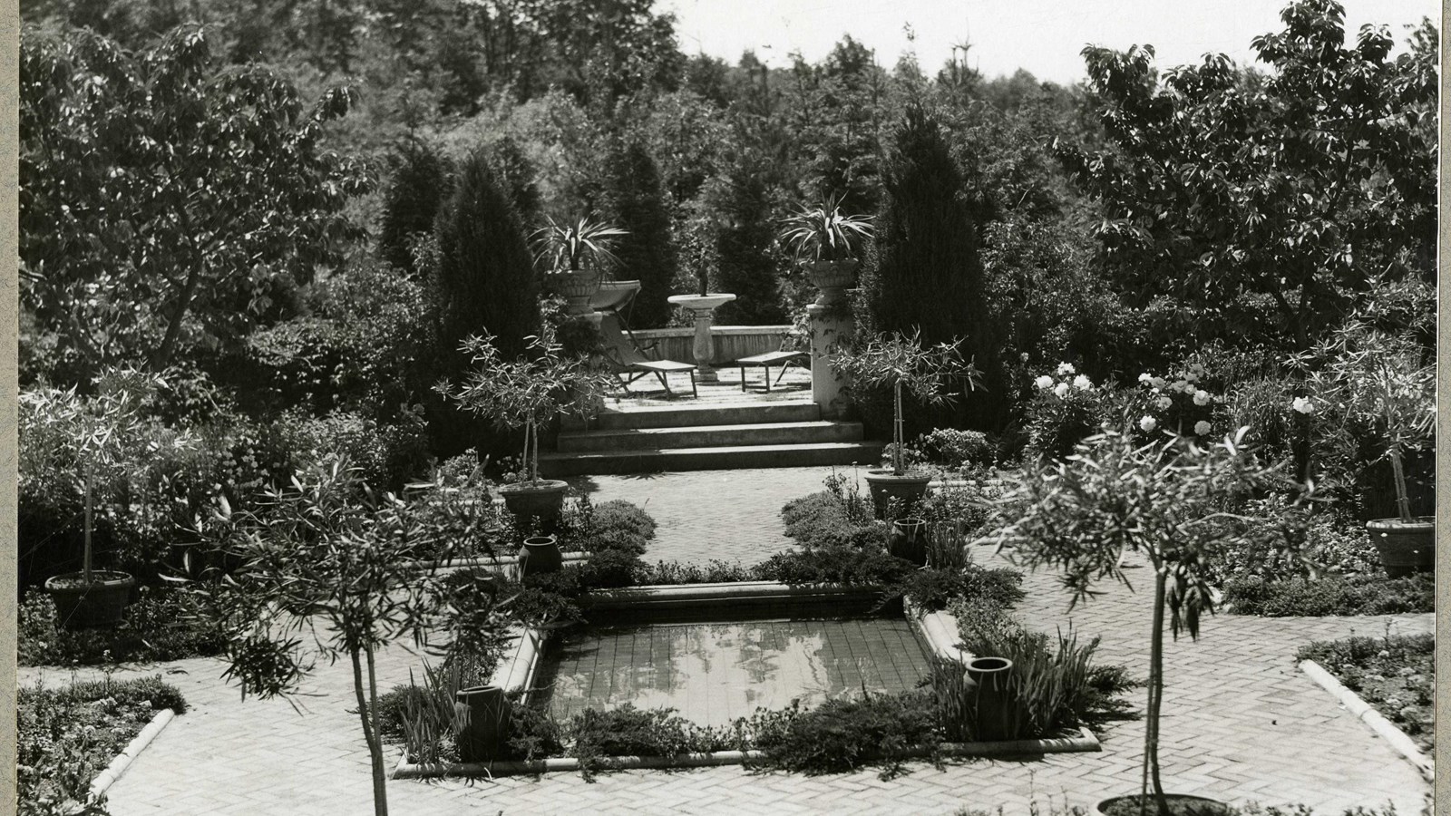 Black and white of sunken garden with rectangular pool, straight lined cement paths lined with trees