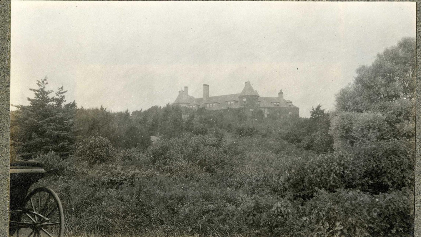 Black and white of hill dense with trees and shrubs, large home on top of hill
