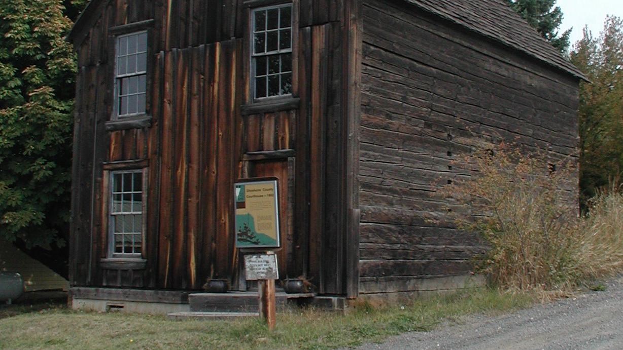 A wooden two story building with coniferous trees on the back side and a gravel road on the right