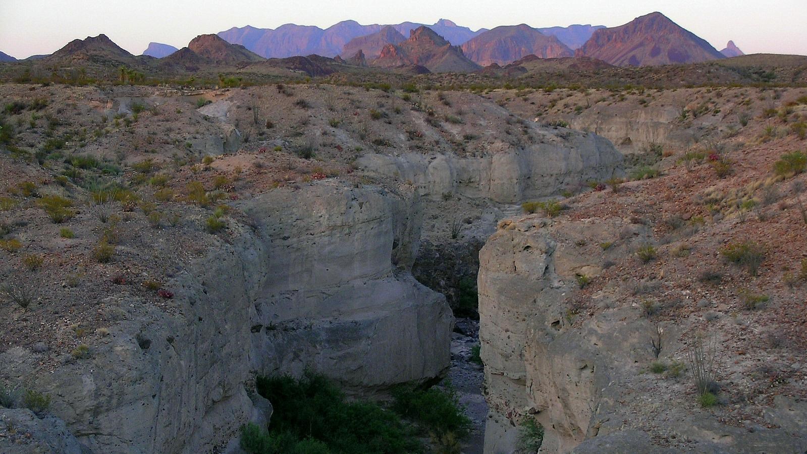 The setting sun casts a glow over a canyon composed of gray tuff, with mountains rising behind.