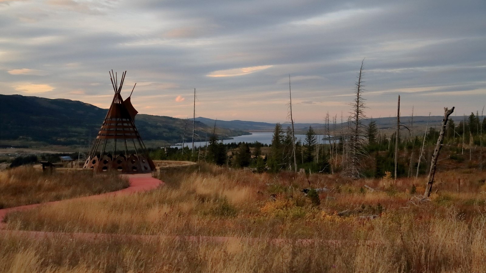 Photo of a grassy area at sunset with mountains and river available in the distance with metal tipi