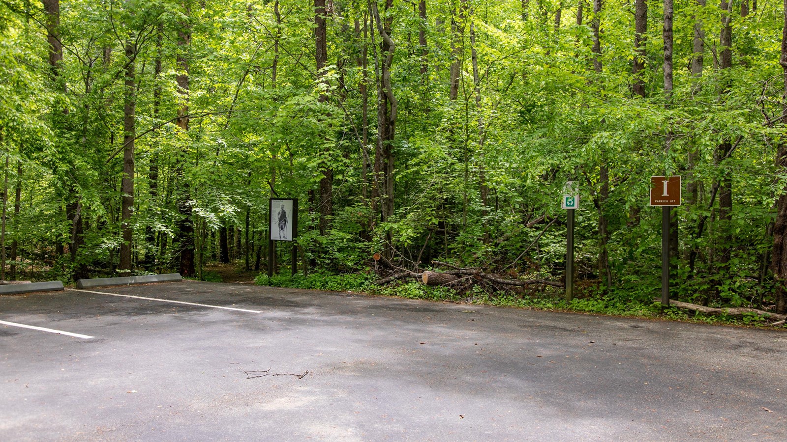 A brown sign with the letter I stands in front of a paved parking lot in the woods.