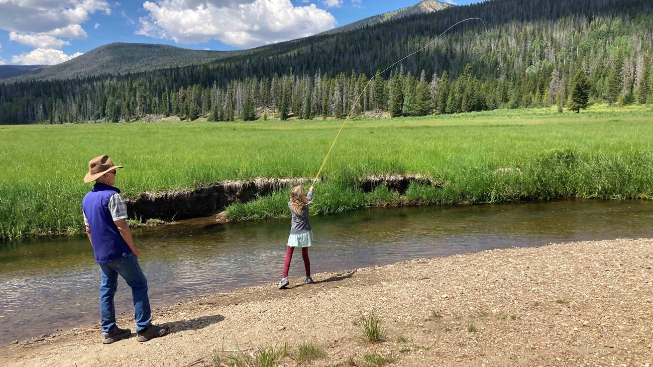 A youth is fishing in a mountain river while an adult supervises