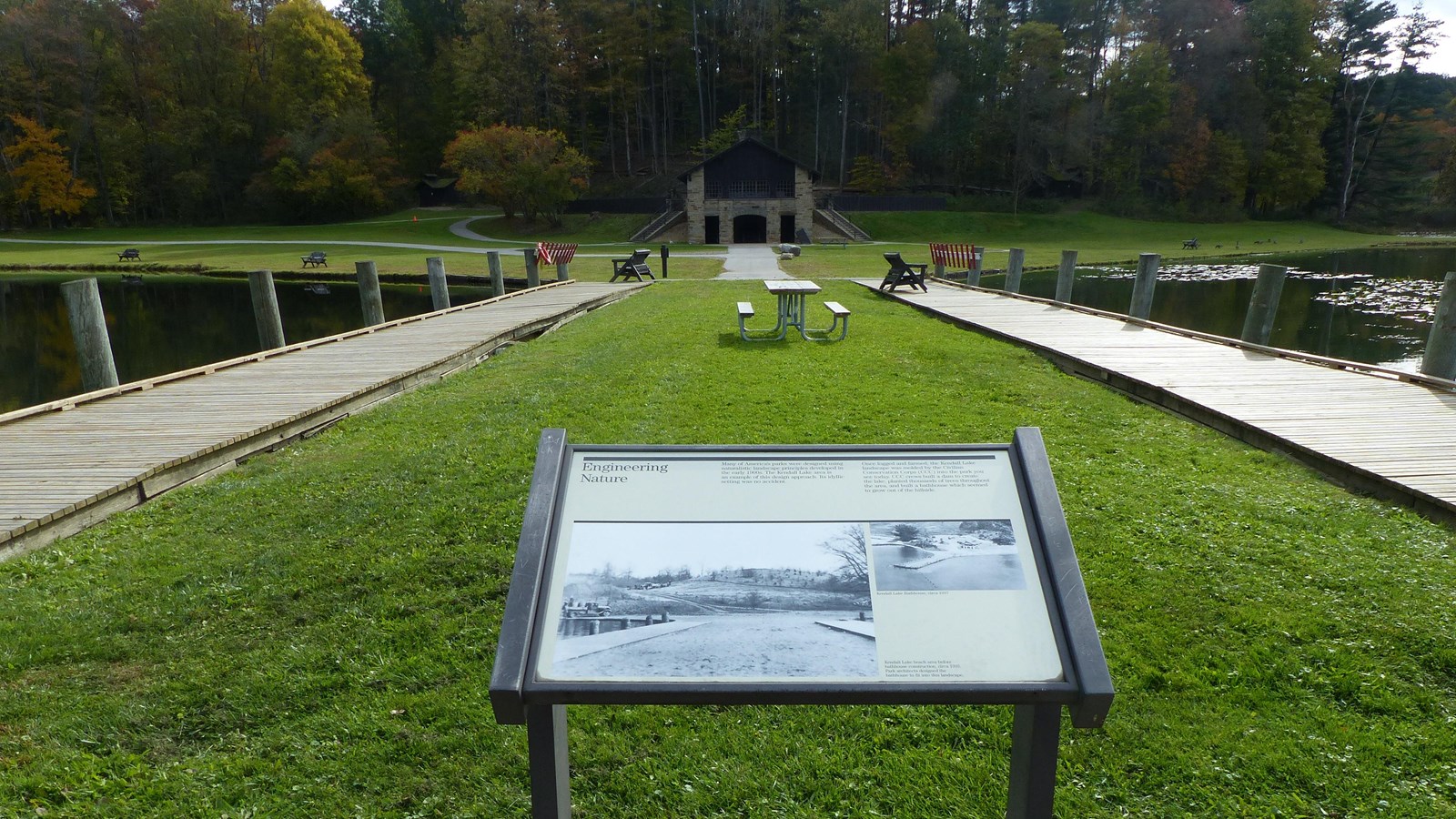 Informational panel on wide grass pier edged by wooden boardwalk; rustic shelter in the distance.