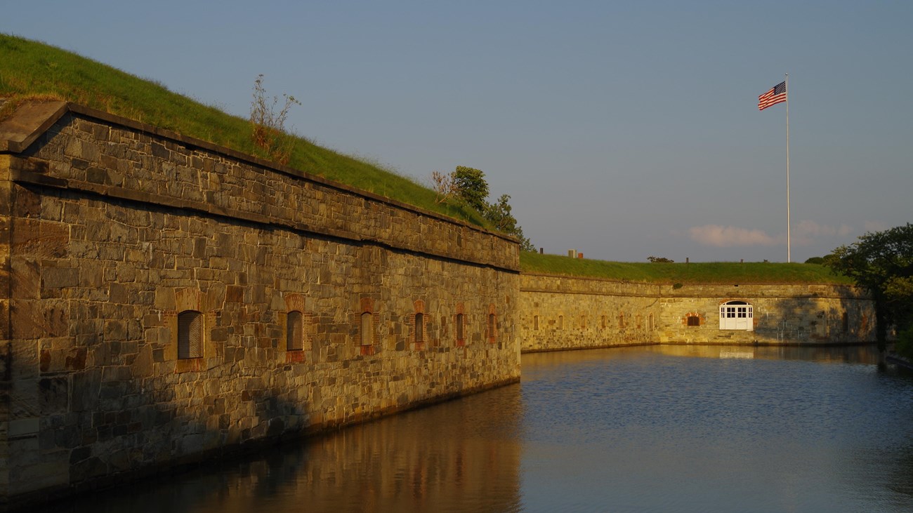 The setting sun lights a stone fort wall where a US flag flies on a high staff. 