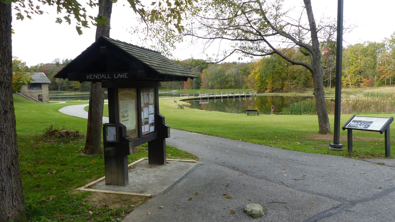 Paved path leads past bulletin board and graphic panel to lake with pier; shelter and woods beyond.