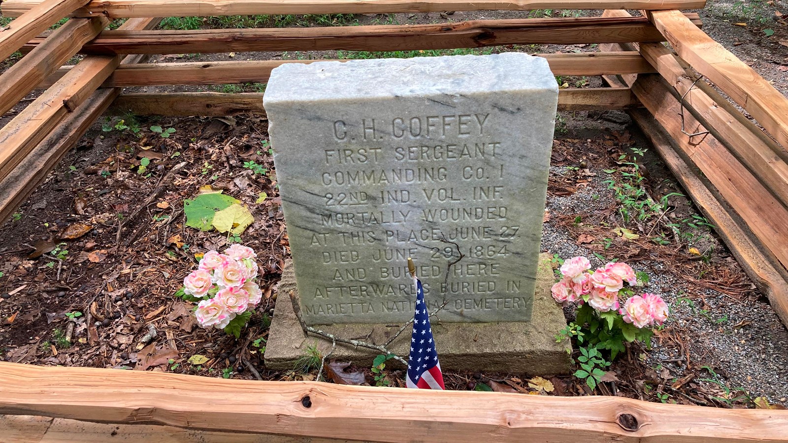 A square stone marker surrounded by a fence and flower arrangements sits near dirt trail. 