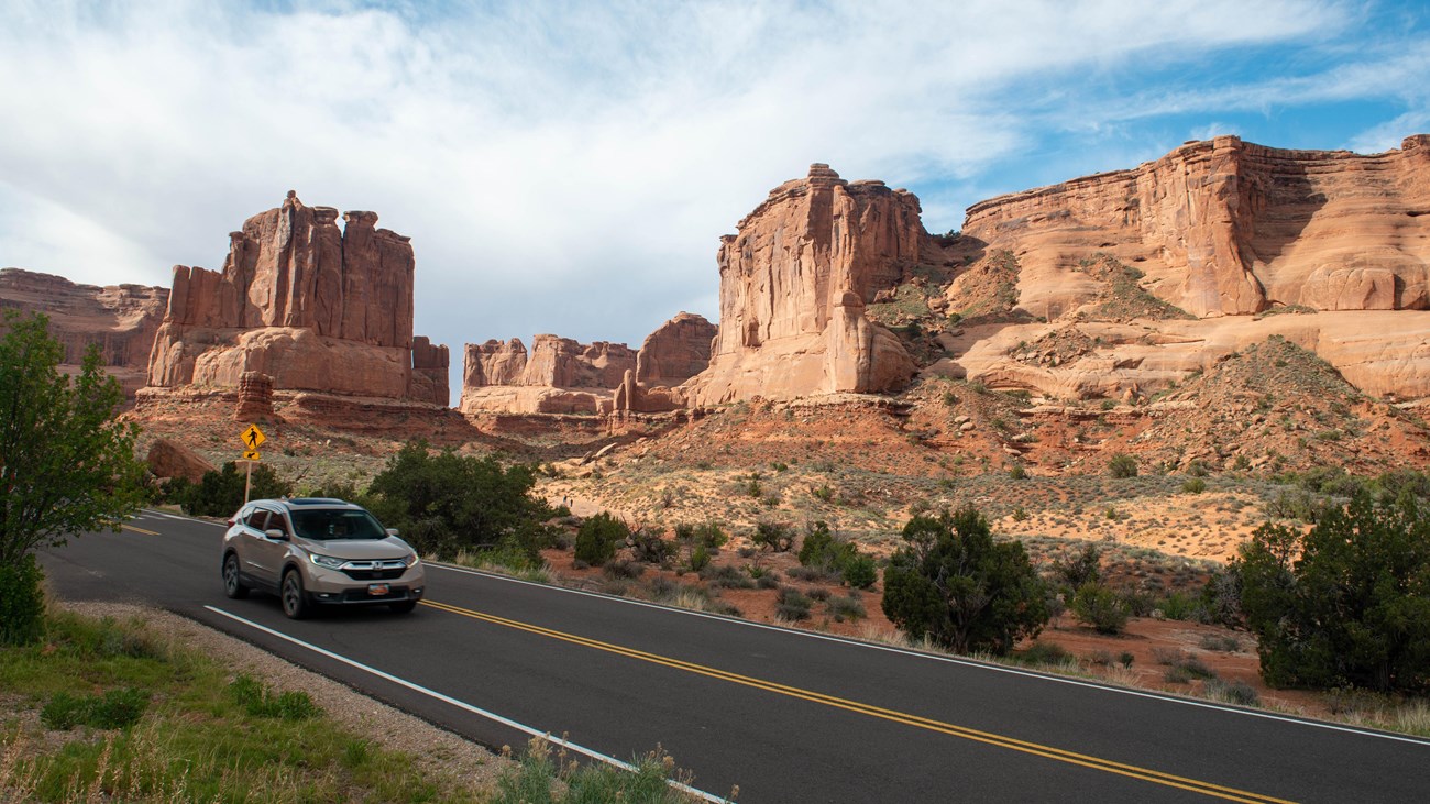 A tan car drives through Arches, sandstone features are visible in the back. 