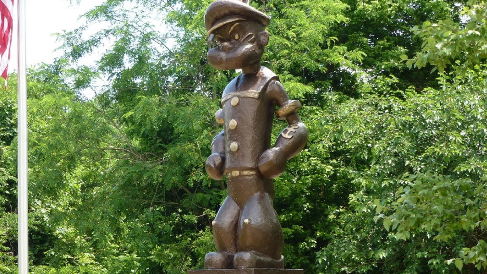 A bronze statue of Popeye the Sailor iin front of green trees